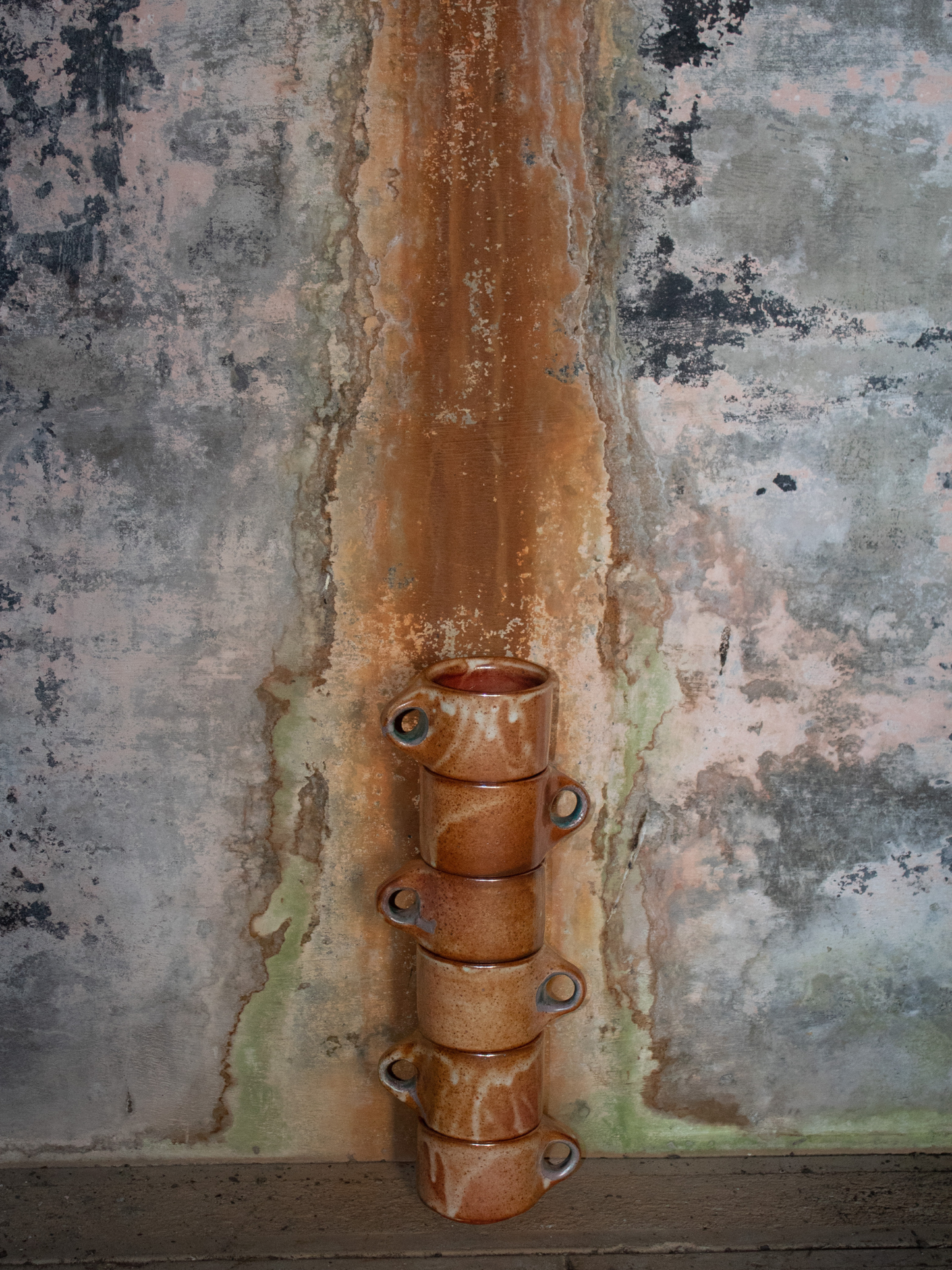 Rust-colored ceramic mugs against a textured wall with visible rust and discoloration.