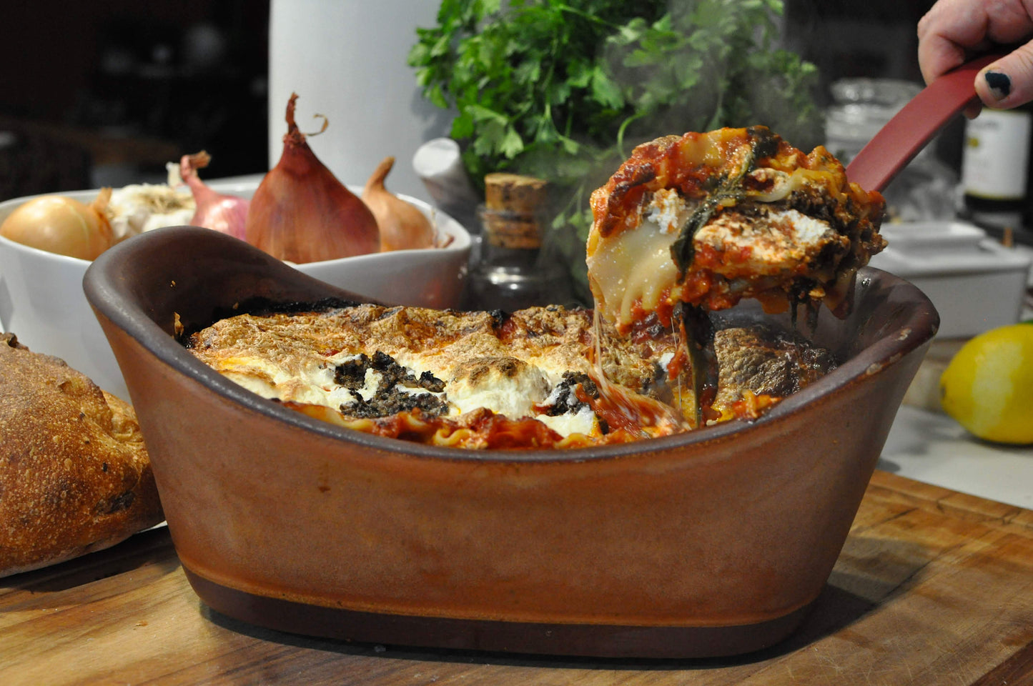Baked dish with cheese and herbs being served with a red spatula, surrounded by bread and vegetables.