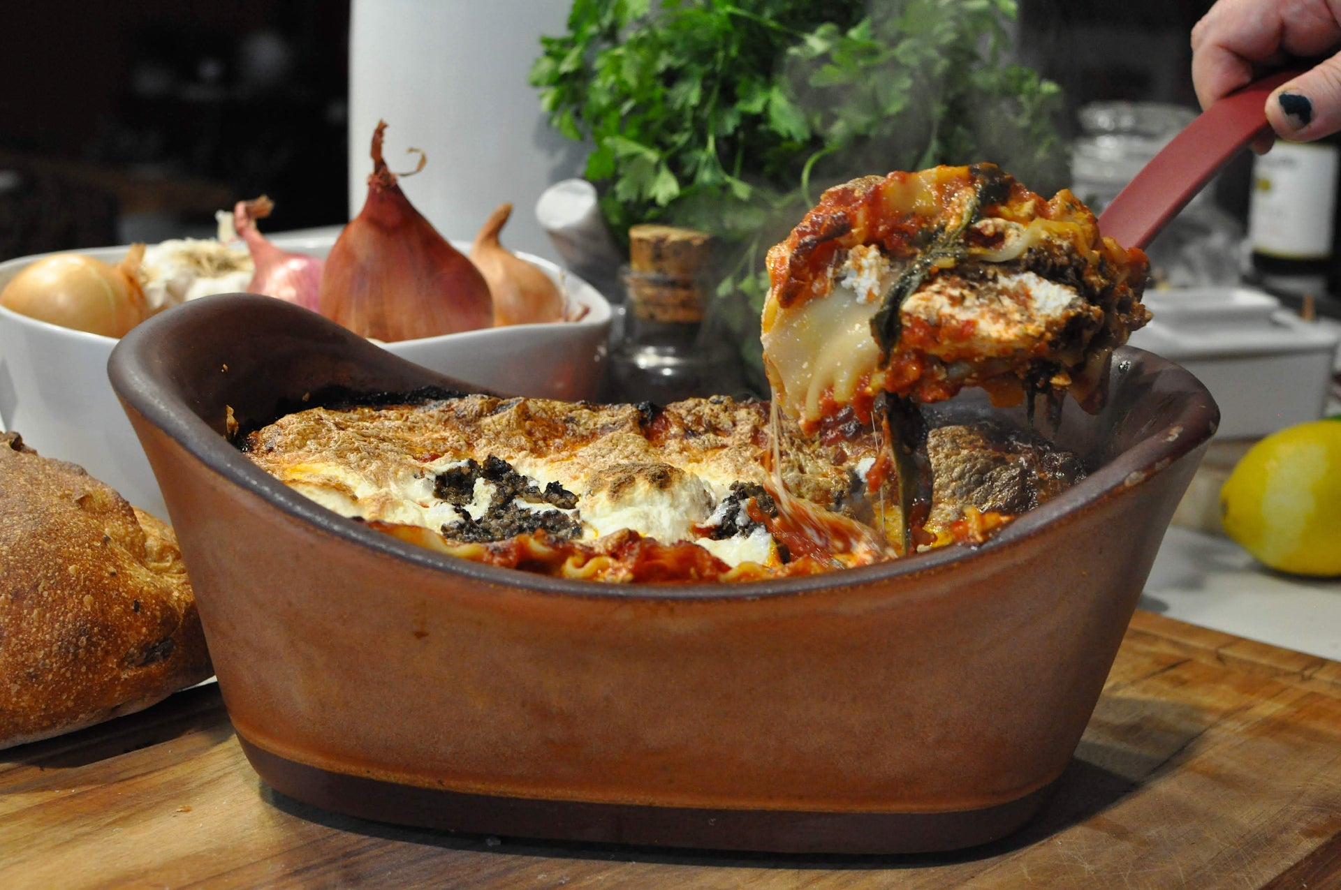 Baked dish with cheese and herbs being served with a red spatula, surrounded by bread and vegetables.