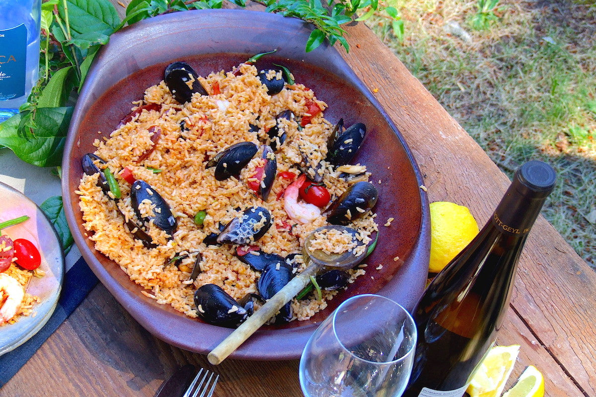 Seafood dish with mussels and rice in a brown bowl on a wooden table with a bottle of wine and lemon slices.