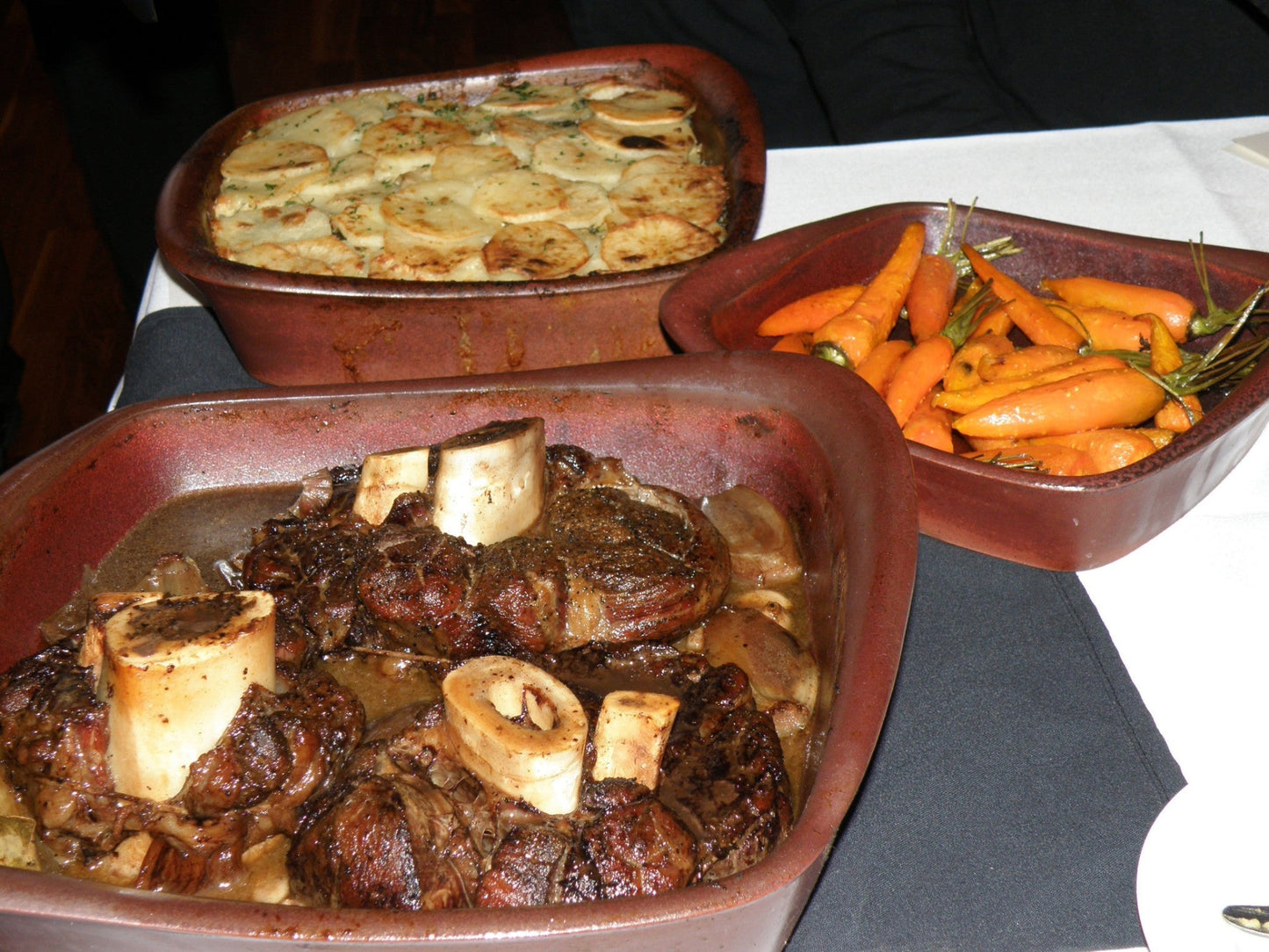 Three ceramic dishes with roasted meats, vegetables, and a gratin dish on a white tablecloth.
