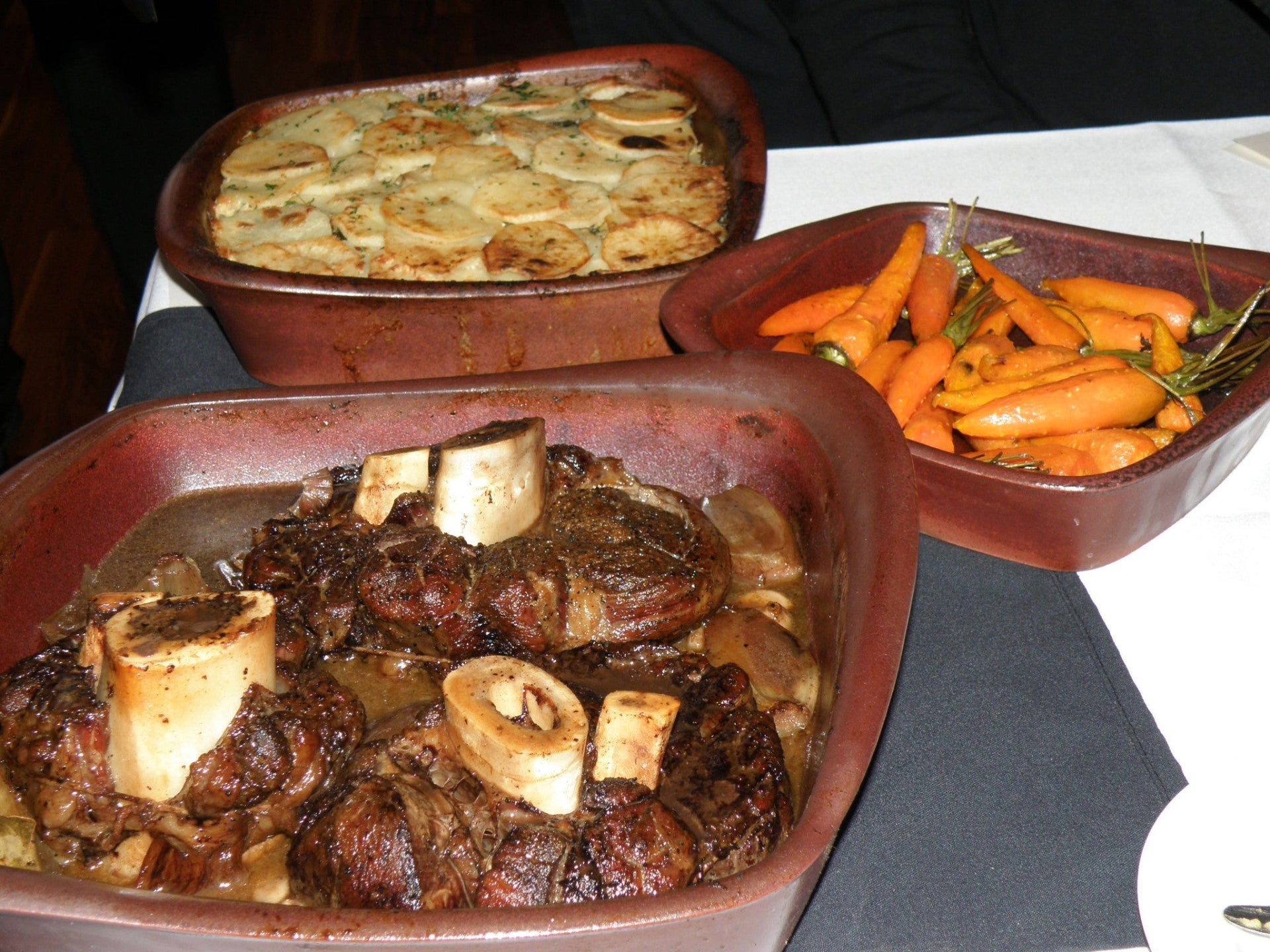 Three ceramic dishes with roasted meats, vegetables, and a gratin dish on a white tablecloth.